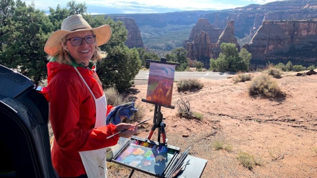 Painter in red jacket and tan hat, painting outside at a canyon overlook.